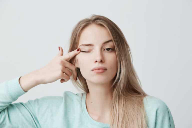 Human face expressions.Thoughtful young beautiful female with blonde dyed straight hair in light blue long-sleeved sweater touching with finger eye, looking at camera with serious and calm expression